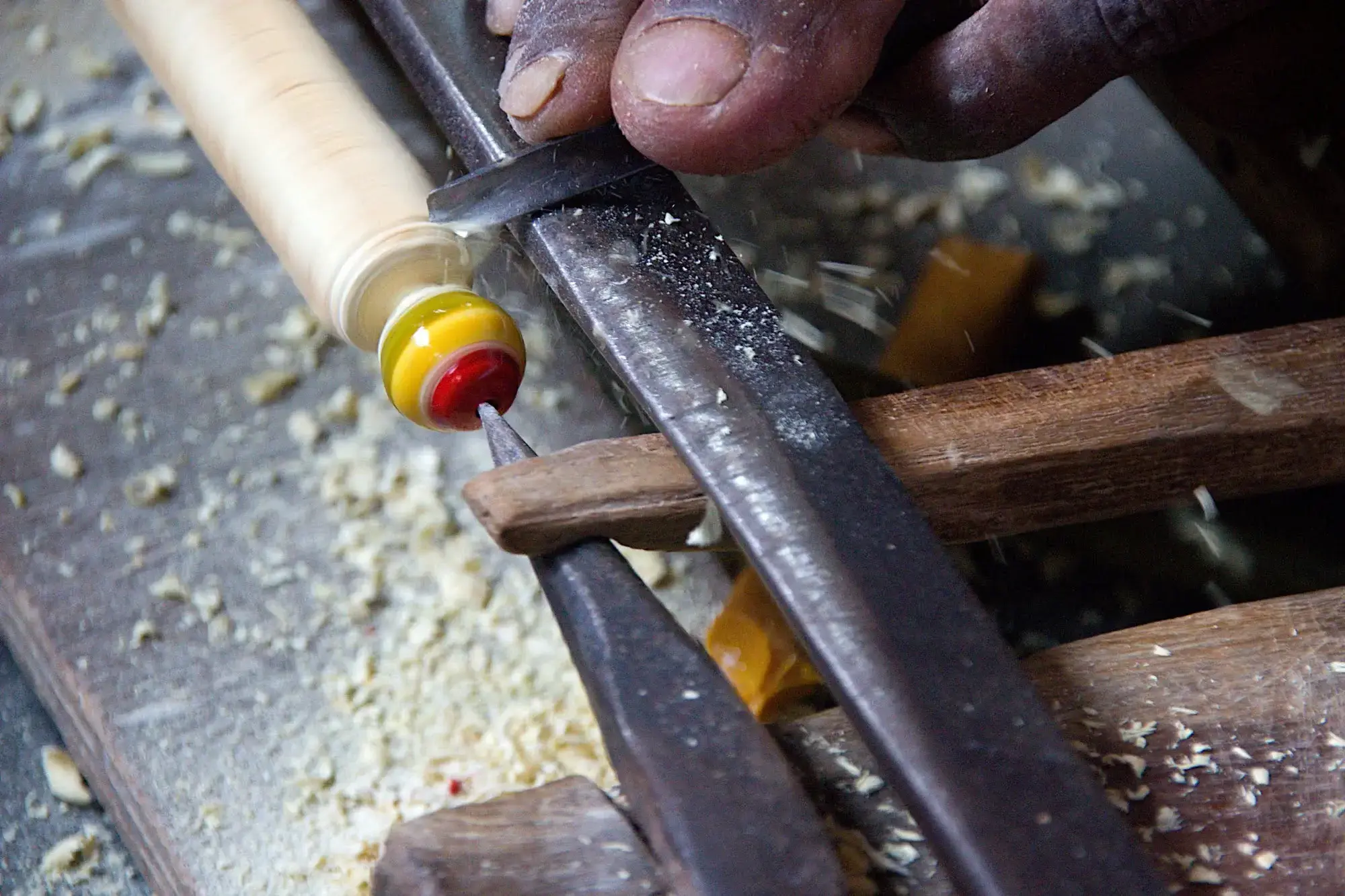 Close-up of a craftsman's hand guiding a chisel along a wooden spindle, creating wood shavings on a workbench.