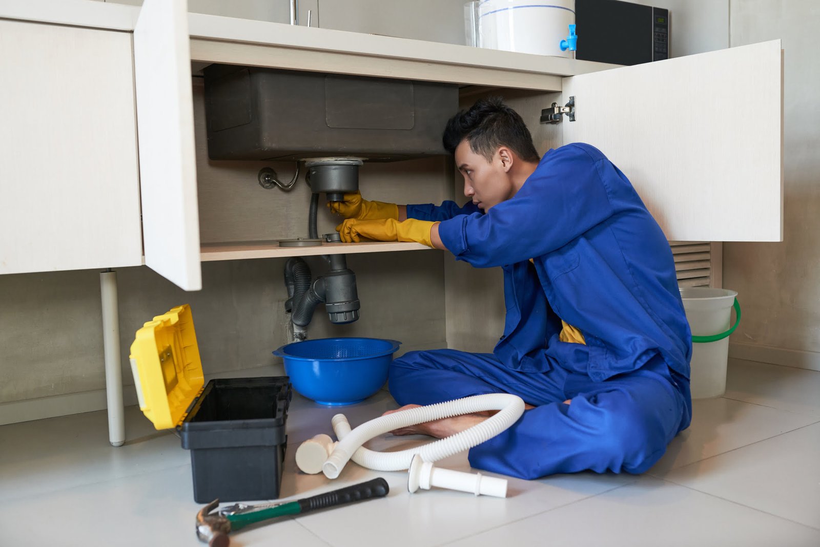 Plumber in blue overalls kneels under a kitchen sink, repairing exposed pipes with yellow gloves nearby a blue bowl and tools.