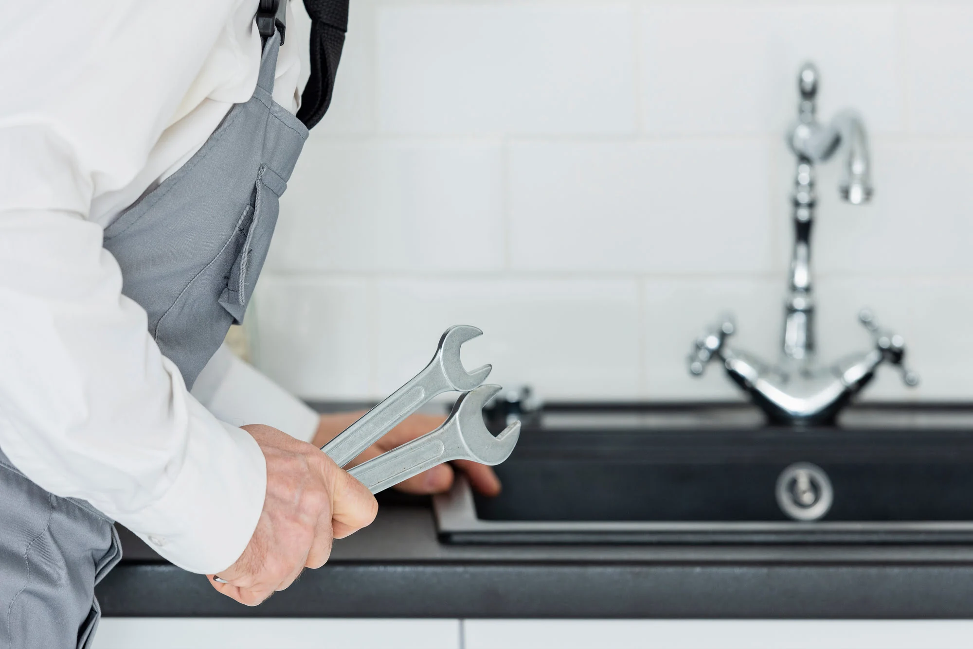 Plumber wearing grey overalls holding two wrenches beside a kitchen sink.
