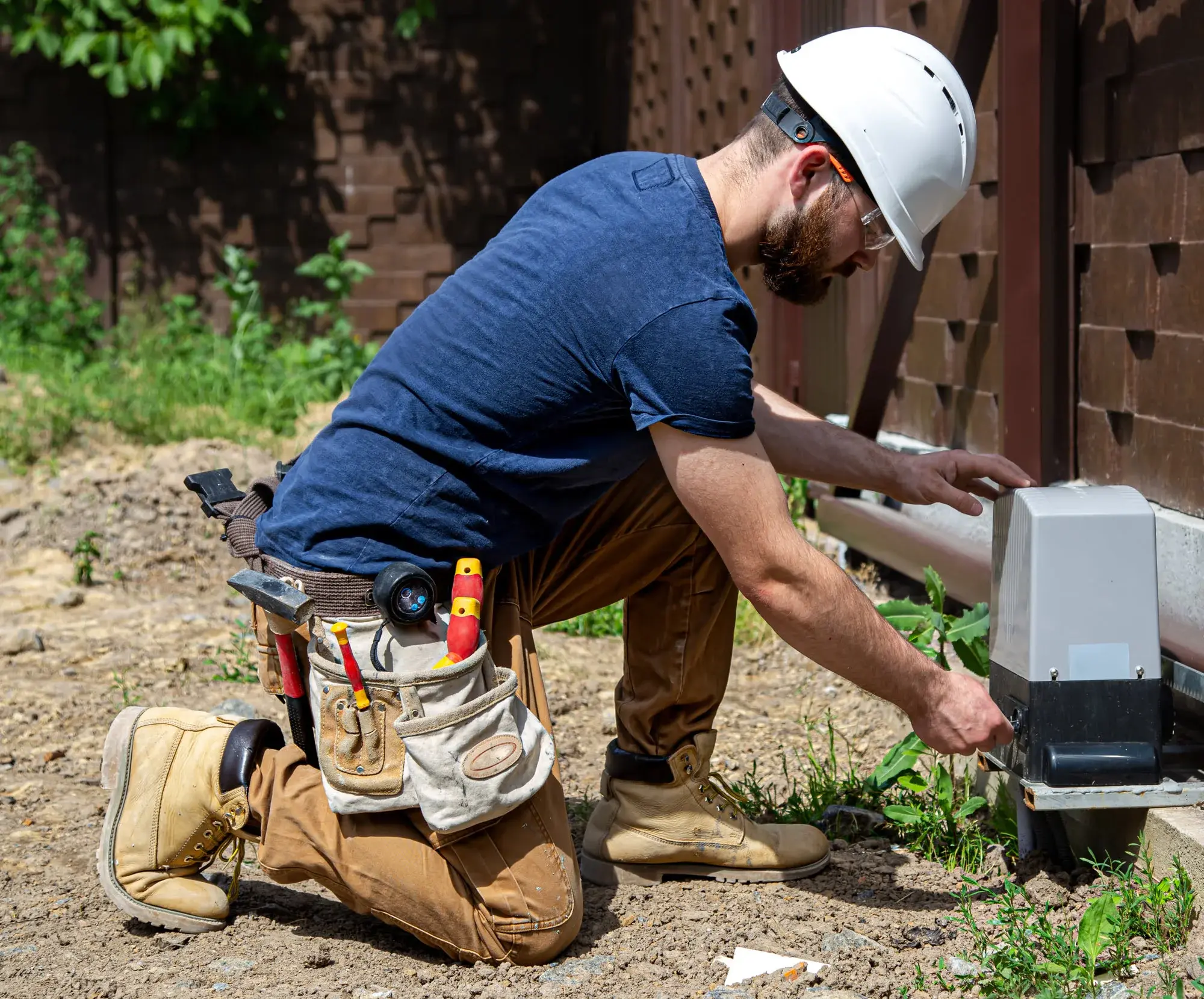 Construction worker kneeling outdoors, wearing a white hard hat and safety glasses, working with a metal utility box.