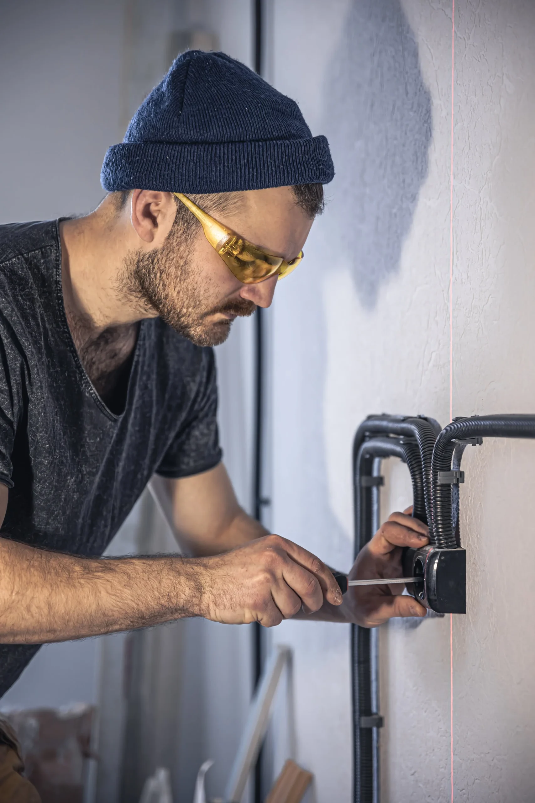 Man in a blue knit beanie and yellow safety glasses tightening a screw on an electrical panel mounted to a wall.