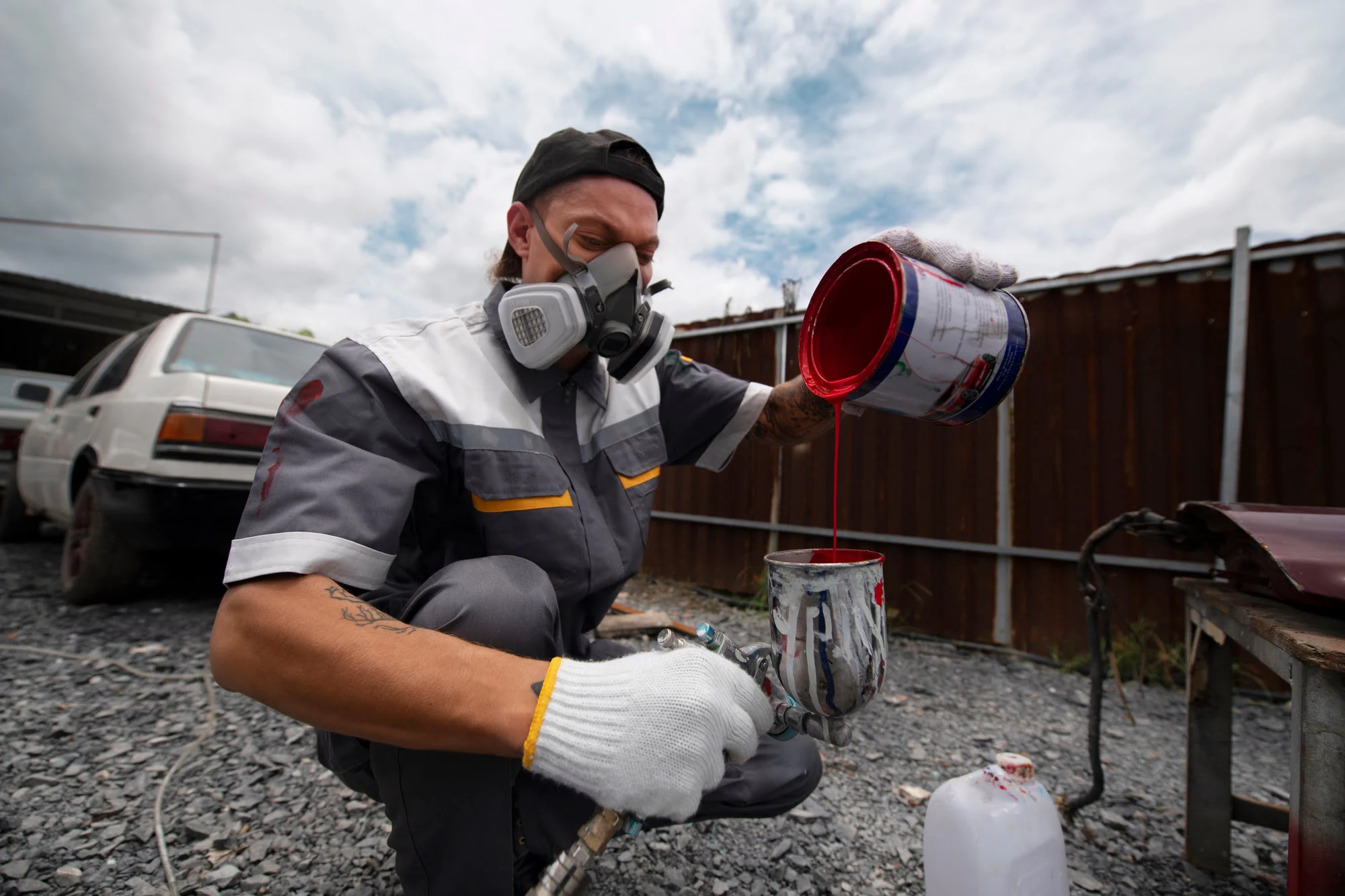 Worker wearing a respirator mask and gloves pours red paint from a can into a container outdoors near a gravel yard.