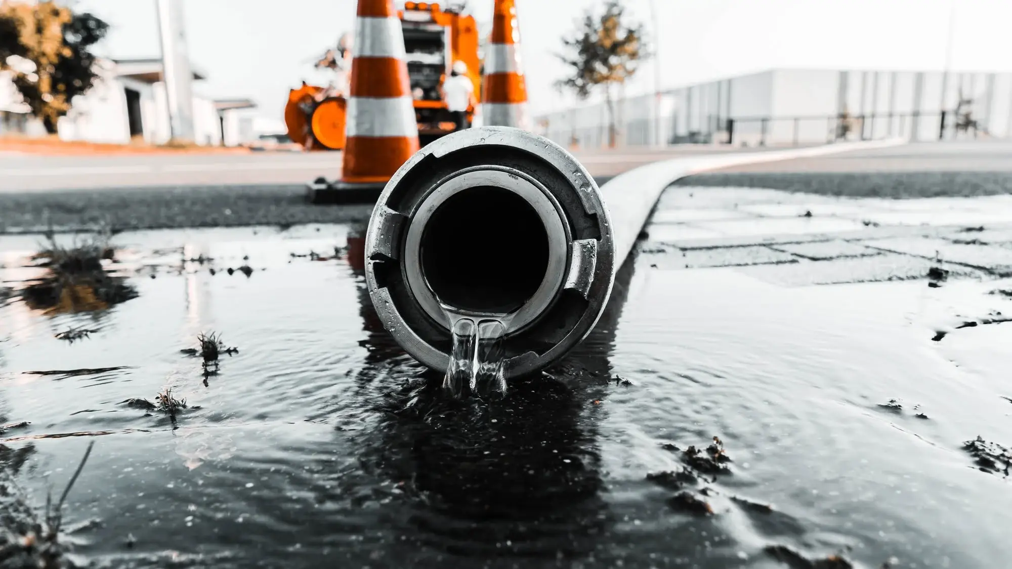 Close-up of a metal pipe outlet spilling water onto a wet construction-site road, orange traffic cones in the blurred background.