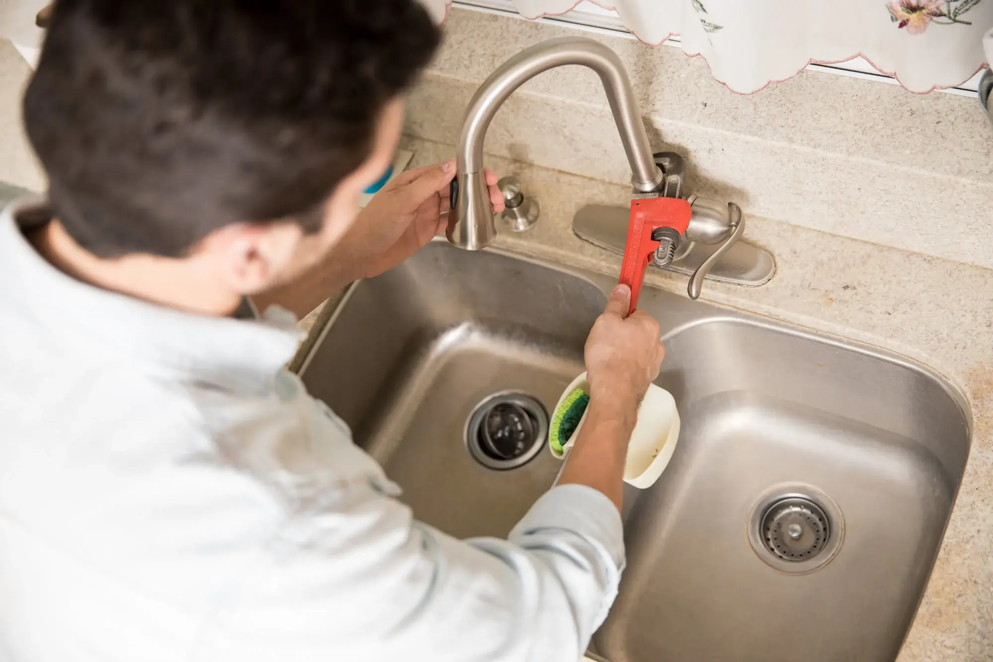 Person using a red pipe wrench to tighten a kitchen faucet at a sink.
