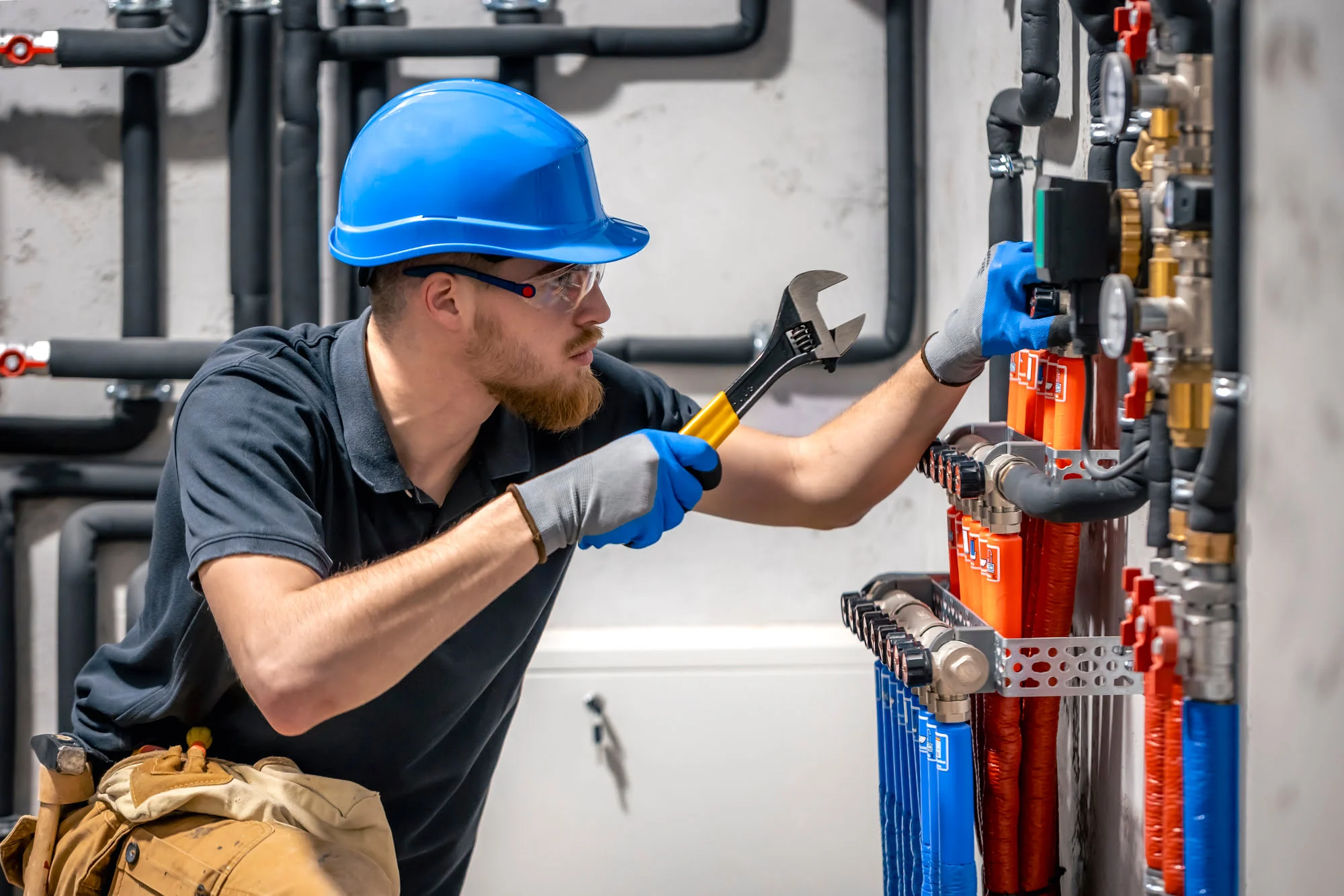 Maintenance worker in a blue hard hat and safety glasses tightens a valve on an industrial control panel with red and blue cables.