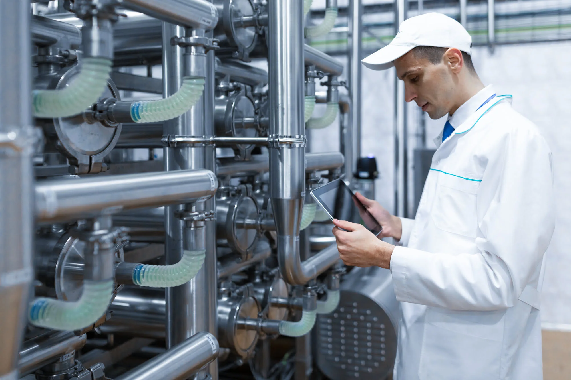 Industrial worker in a white lab coat and cap inspecting complex stainless steel machinery while holding a tablet.