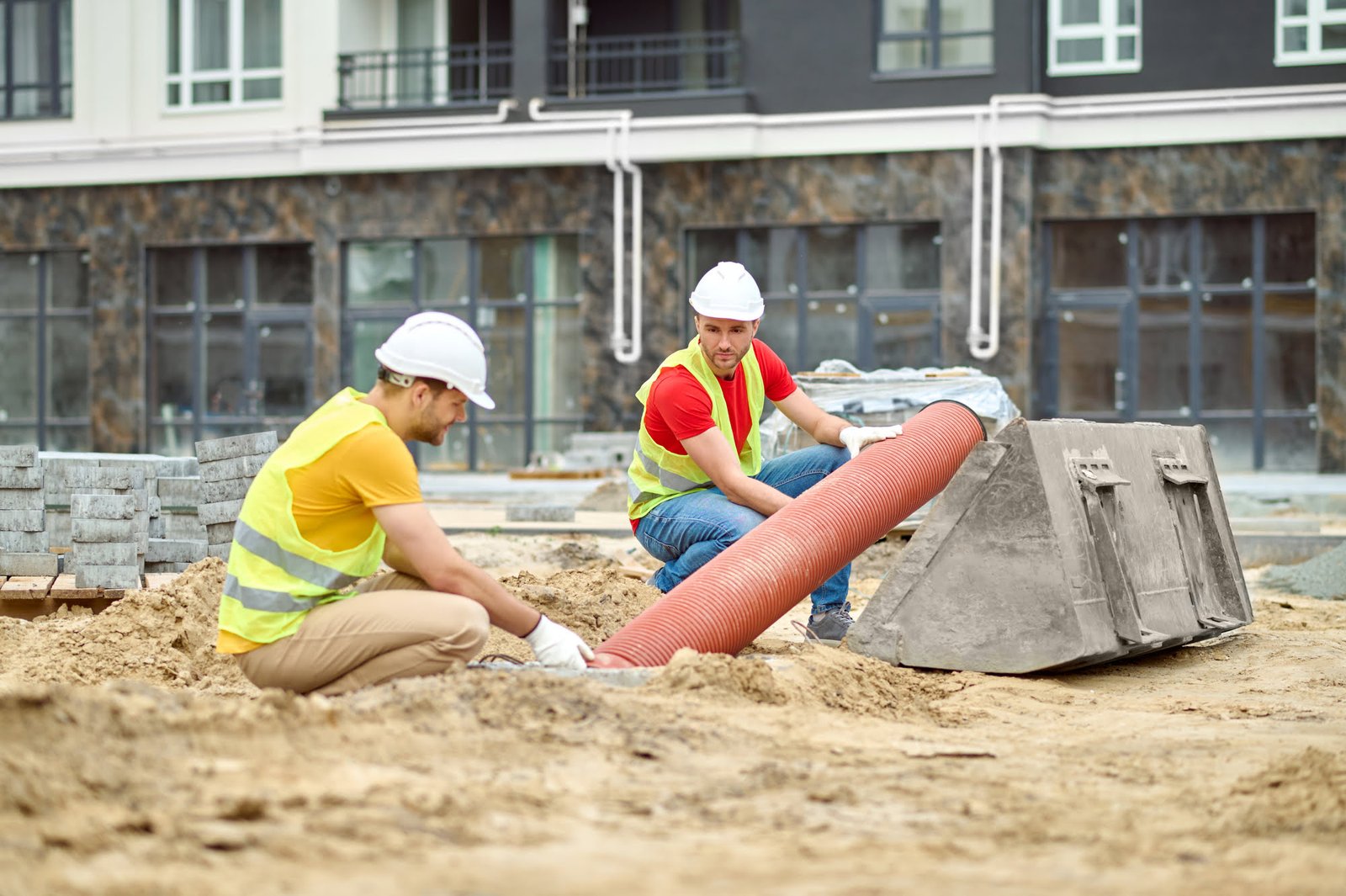 Two construction workers in hard hats and high‑visibility vests lay a large orange pipe on a dirt construction site with a building under construction in the background.