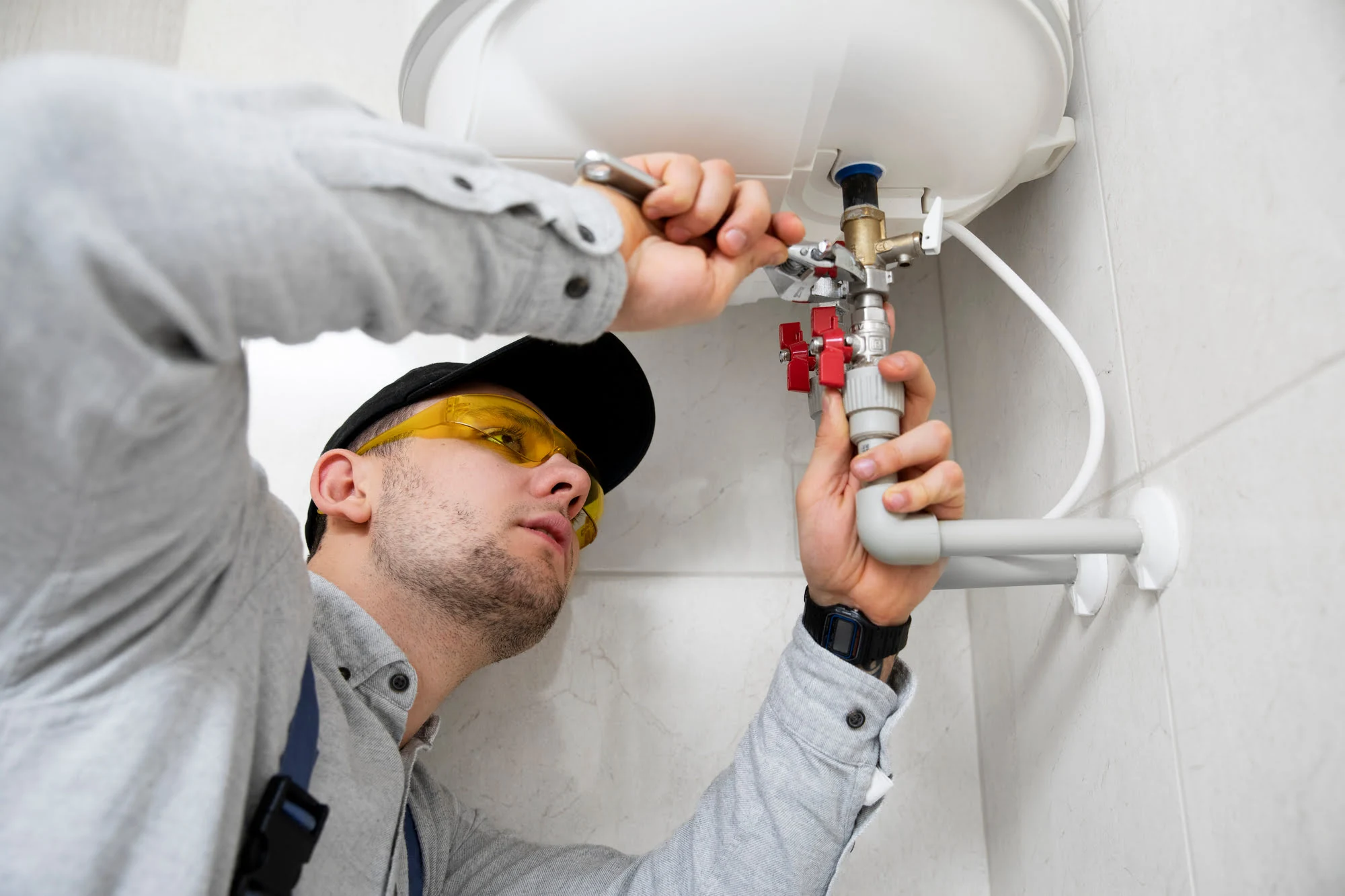 Plumber tightening a valve under a water heater with safety glasses and a gray shirt.